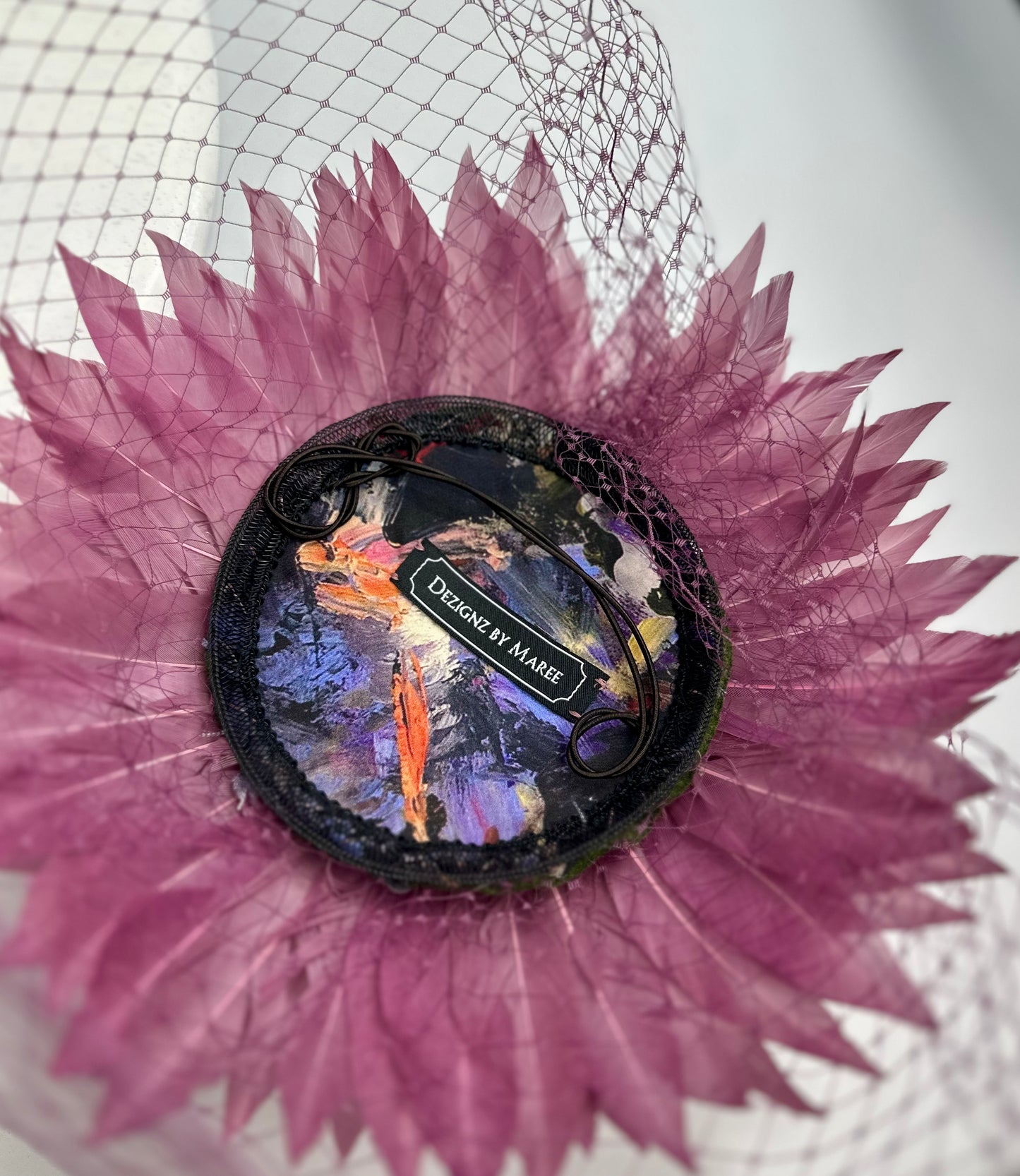 Underneath of hat headpiece with elastic, purple feathers, purple veil netting and 'Dezignz by Maree' label on a white background