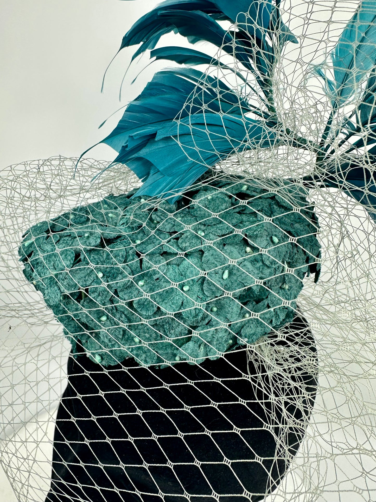 Abstract headpiece with teal box hat, blue teal feathers, white silver netting veil on white background