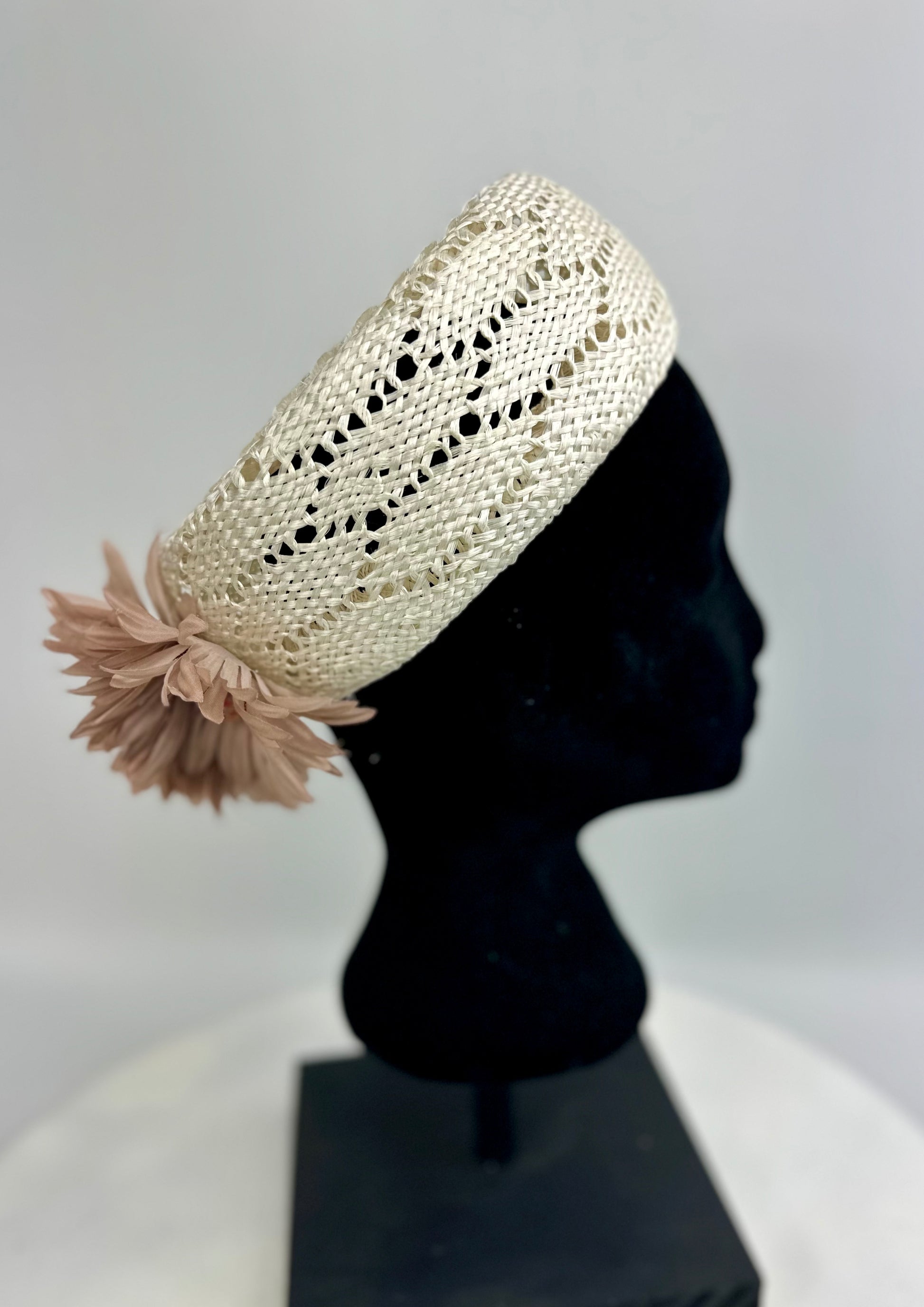Cream-coloured woven pillbox hat with dusty rose beige floral design on back, on a black mannequin head against a gray background