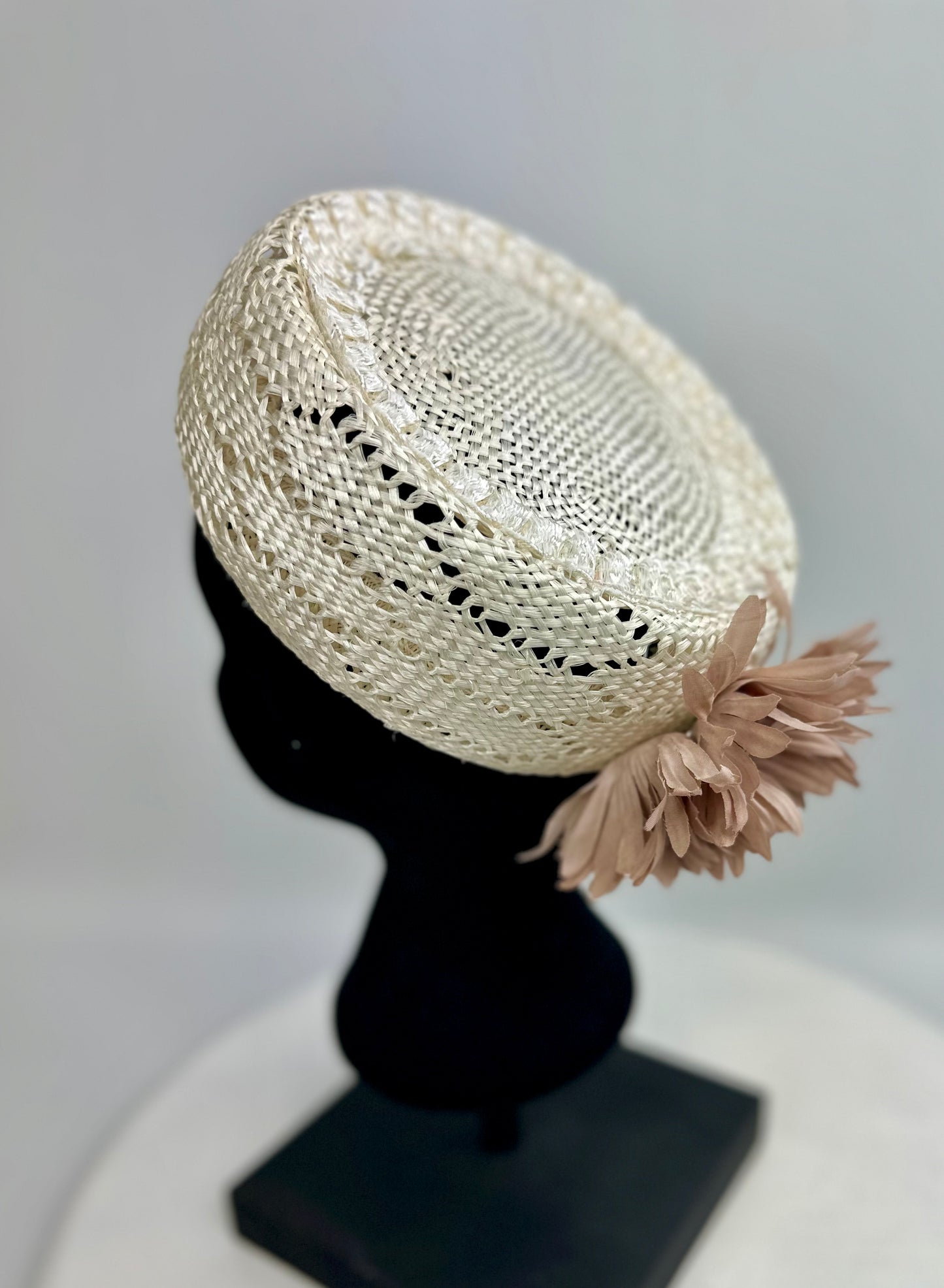 Cream-coloured woven pillbox hat with dusty rose beige floral design on back, on a black mannequin head against a gray background