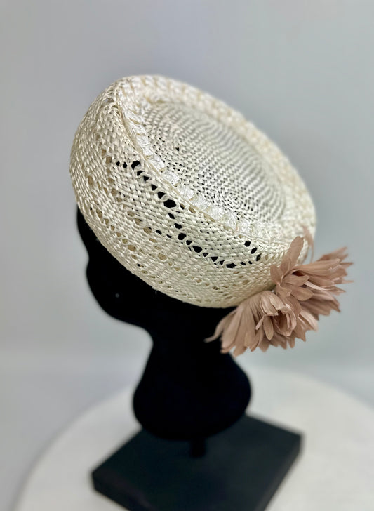 Cream-coloured woven pillbox hat with dusty rose beige floral design on back, on a black mannequin head against a gray background