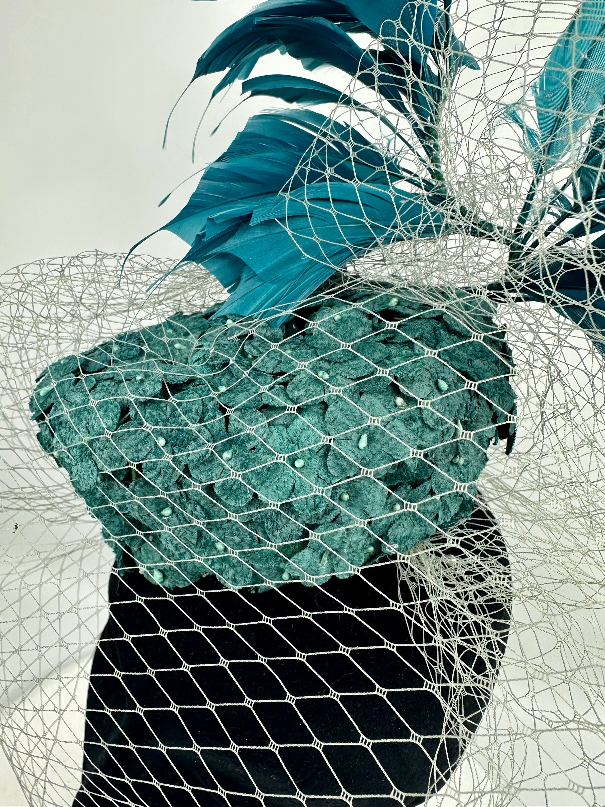 Abstract headpiece with teal box hat, blue teal feathers, white silver netting veil on white background