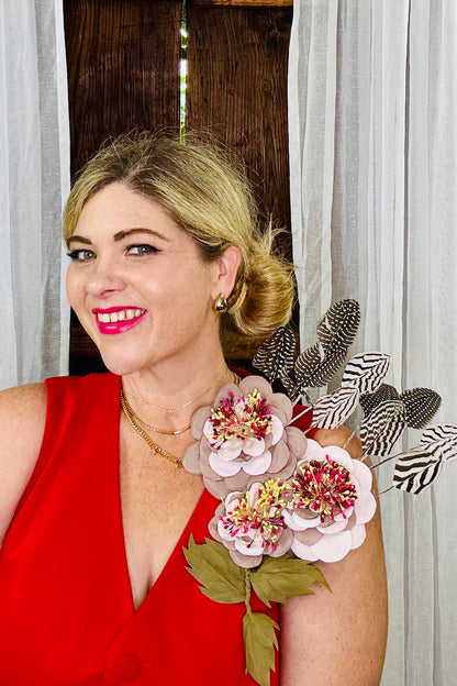 Woman wearing a red dress with decorative dusty rose and pastel pink floral and feather accessory brooch with maroon, pink and yellow details against a neutral background