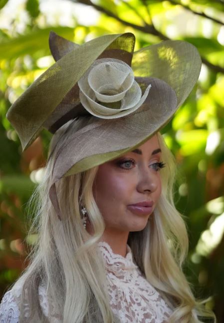 Woman wearing Sueellen hat headpiece, a large decorative hat with floral details against a blurred green background