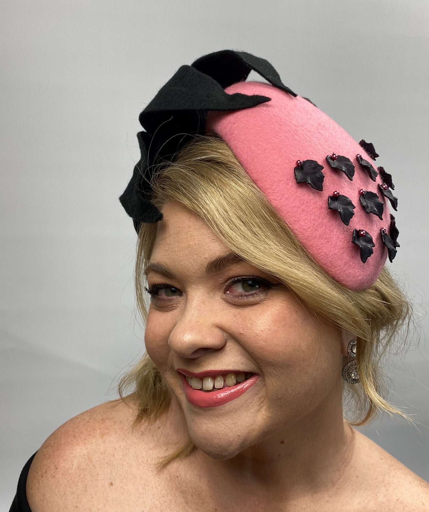 A woman wearing a pink felt race day hat with a large black felt flower and black leaf findings, attached to a black headband.