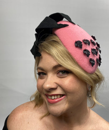 A woman wearing a pink felt race day hat with a large black felt flower and black leaf findings, attached to a black headband.